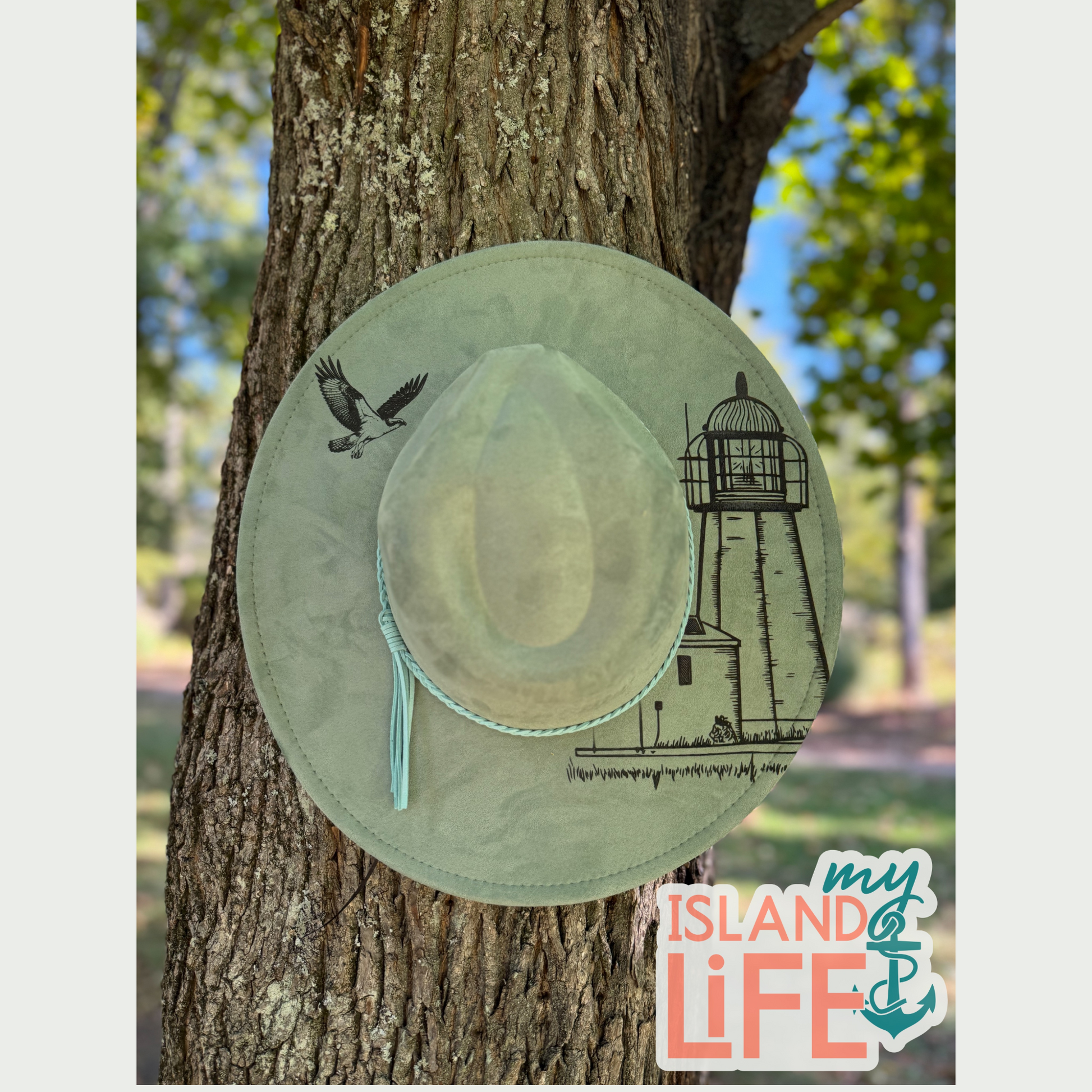 Light green hat with lighthouse nature-themed design hanging on a tree trunk, with 'My Island Life' logo in the corner.