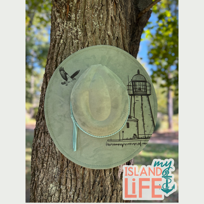 Light green hat with lighthouse nature-themed design hanging on a tree trunk, with 'My Island Life' logo in the corner.